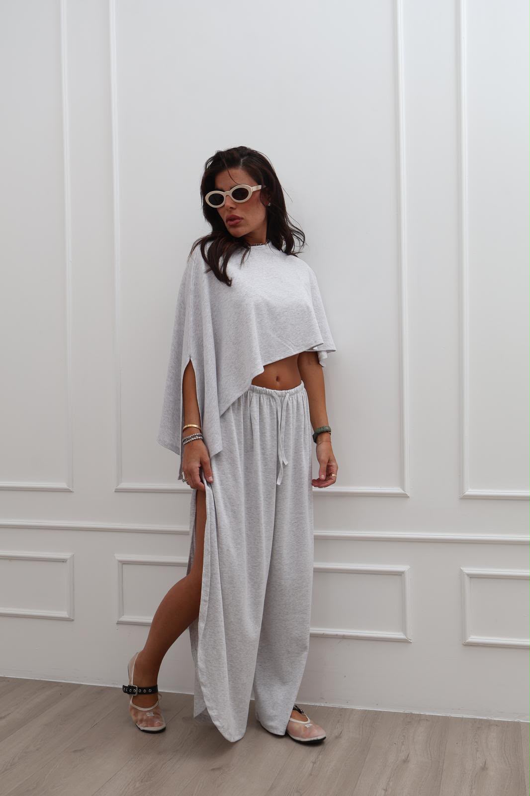 A woman modeling a light gray two-piece lounge set consisting of a cropped tunic top and matching wide-leg drawstring pants, standing against a white paneled wall.